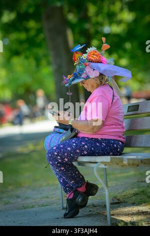 Woman sitting on park bench wearing ornately flowery, decorated hat at music festival Stock Photo