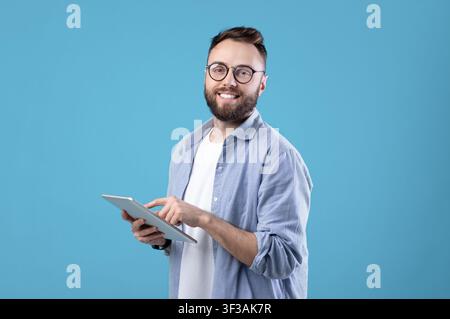 Positive young guy in casual wear using tablet computer over blue studio background Stock Photo