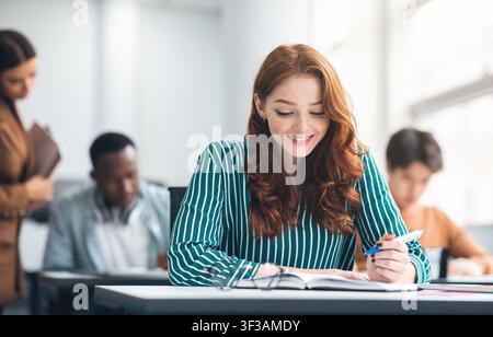 Smiling lady sitting at desk in classroom writing in notebook Stock Photo