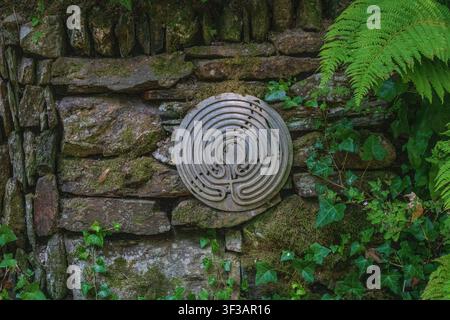 A patterned stone labyrinth carved into a round disc, fixed to a weathered, moss-covered stone wall, surrounded by lush green ivy and fern foliage. Stock Photo