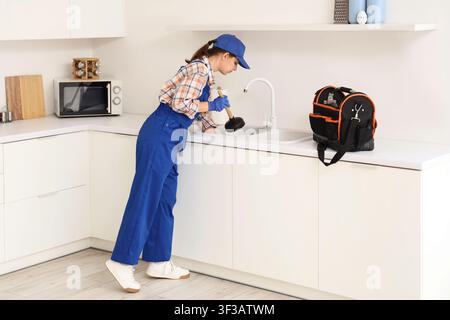 Young female plumber in blue uniform and cap using a plunger to clear a clogged kitchen sink, She wears protective gloves while performing the repair Stock Photo