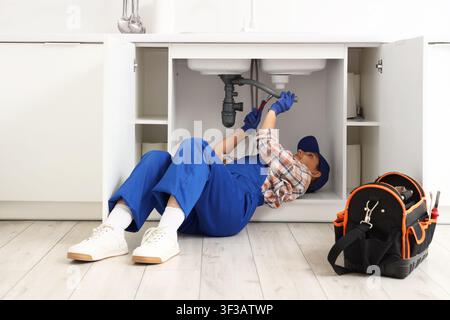 Female plumber in blue uniform and cap repairing a kitchen sink pipe with a wrench, Home maintenance and repair service Stock Photo