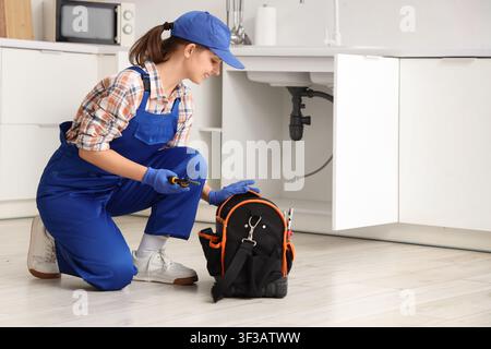 Female plumber in blue uniform and cap fixing a kitchen sink with tools from her bag, She is using tools from her open toolbox, demonstrating professi Stock Photo