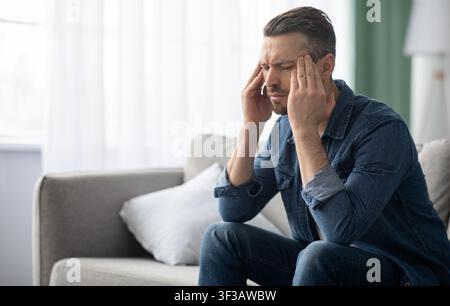 Stressed man having headache, sitting on sofa at home Stock Photo
