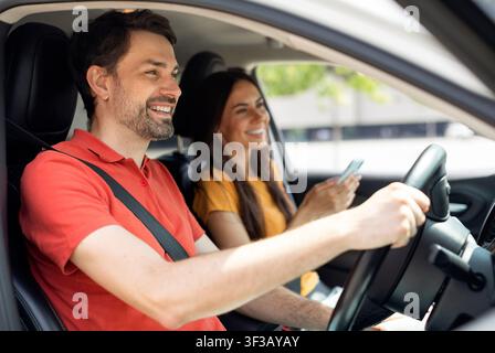smiling millennial couple sitting inside luxury car, woman holding smartphone Stock Photo