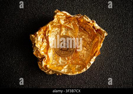 Physical Bitcoin token resting on crumpled gold foil against a textured black background, symbolizing digital wealth Stock Photo