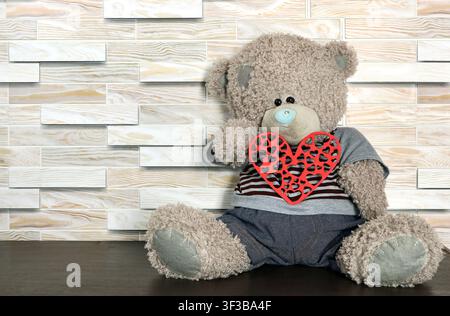 A teddy bear holds a red plywood heart in its paws. A toy bear sits on a wooden shelf. Stock Photo