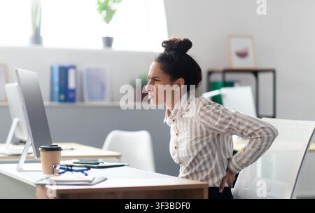 Young businesswoman having acute back pain, massaging aching muscles, sitting at desk in office interior, copy space Stock Photo