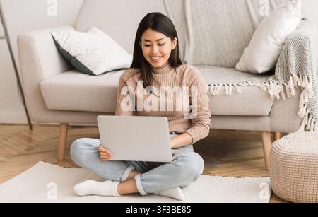 Asian woman networking on laptop, sitting on floor Stock Photo