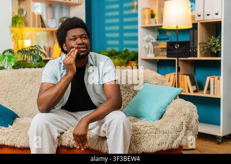 African American young man on home sofa touches cheek with toothache pain showing worried face in living room. Black guy on home sofa feels dental problem with discomfort needing dentist care at home. Stock Photo