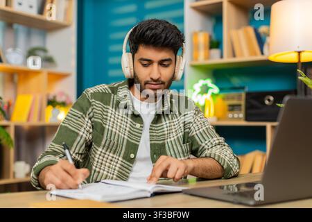 Indian man on home table wears headphones listening to lesson taking notes with serious focus. Arabian guy on home table nods during remote class or client call using laptop notebook for web training. Stock Photo