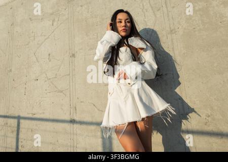 A stylish young Asian woman in a white denim outfit poses confidently against a sunlit textured concrete wall, featuring strong shadows and a low-angl Stock Photo