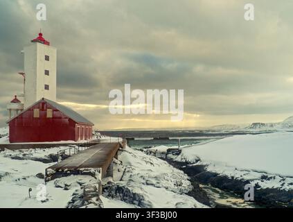 Berlevåg, Norway - 31.03.2019: Red wooden structure and white lighthouse tower standing on snow-draped rocky shoreline under moody sky Stock Photo