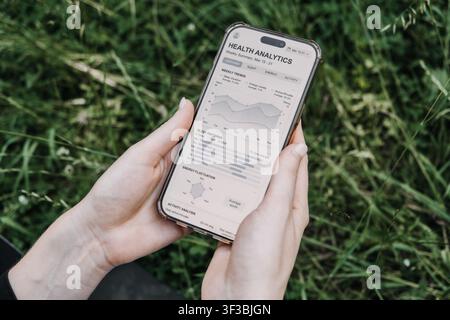 Two hands hold smartphone displaying health analytics app against green grassy background. Mobile health, digital wellness, self tracking, biohacking. Stock Photo
