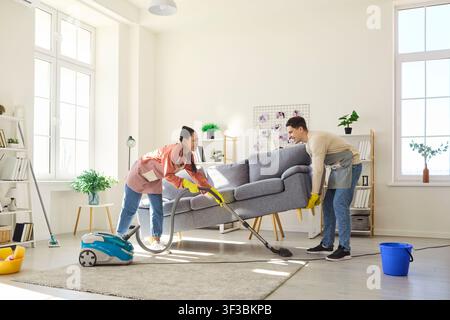 Couple vacuuming under sofa while lifting couch in bright modern living room Stock Photo