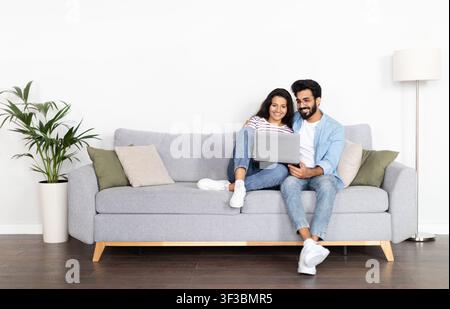 Loving eastern couple sitting on couch at home, using laptop Stock Photo