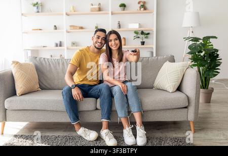Joyful diverse couple watching TV program, sitting on couch at home, full length portrait Stock Photo