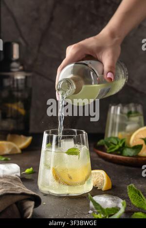 Fresh mojito cocktail and ingredients on dark table background. Pouring soda into a glass with ice cubes, making drink process. Vertical Stock Photo