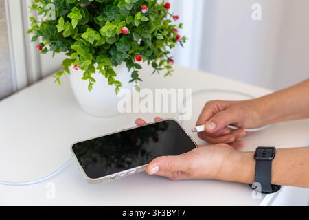 Hands Connecting a Charging Cable to a Smartphone. Hands connecting a charging cable to a smartphone on a white desk beside a potted plant. Stock Photo