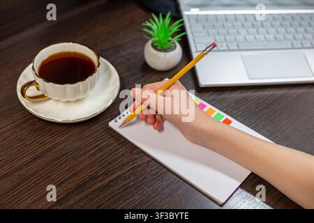 Hand Writing in Notebook Beside Laptop and Tea. Hand holding a pencil over a blank spiral notebook beside tea, a laptop and a small plant on a wooden Stock Photo