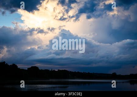 A bright cloudburst lights the sky over a calm lake while dark banks of cloud sweep in from the left and the shoreline rests in silhouette reflecting Stock Photo
