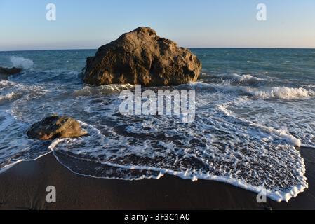 Sea stack on a dark sand shore with incoming white waves at sunset, Cyprus Stock Photo