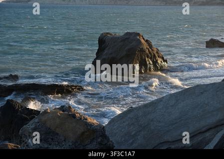 Sea rock surrounded by crashing sea foam on a sandy beach shore at golden hour, Cyprus Stock Photo