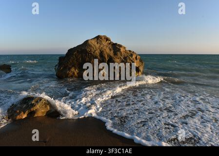 Sea rock surrounded by crashing sea foam on a sandy beach shore at golden hour, Cyprus Stock Photo