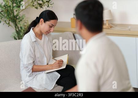 Asian female therapist taking notes during counseling session with male client Stock Photo