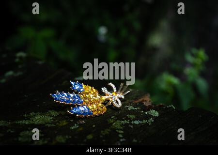 Jeweled bouquet brooch in blue and yellow rests on aged wood with moss. Low light sparkles on crystals while dark greens surround the ornate piece giv Stock Photo