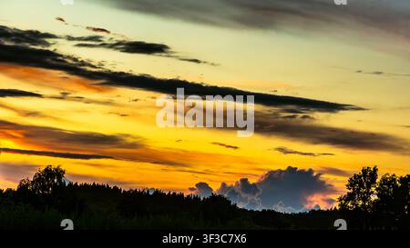Dramatic golden sunset above a dark forest horizon beneath layered evening clouds Stock Photo