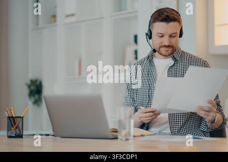 Male customer support agent wearing headset reviews documents beside laptop in home office Stock Photo