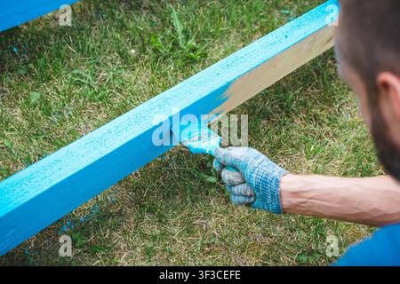 Man painting a wooden plank bright blue outdoors on green grass, wearing protective gloves, concept of renovation, craftsmanship, and DIY home improvement Stock Photo