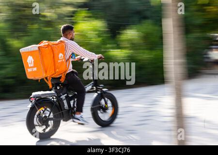 A food delivery rider speeds along a tree-lined path on an electric bike, a large orange thermal bag strapped to his back, captured in motion blur Stock Photo