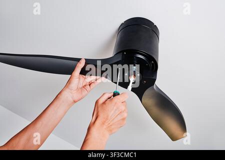 while installing a black three-blade ceiling fan, a man tightens a screw while supporting one blade, with the central fitting exposed against a plain Stock Photo