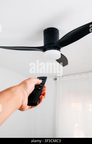 a black three blade ceiling fan with integrated white light is operated by a man using a handheld remote control beneath a plain white ceiling in a br Stock Photo