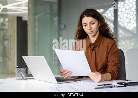 Young businesswoman with long curly hair working diligently, reviewing financial documents and using her laptop at a white desk in a contemporary corporate environment Stock Photo