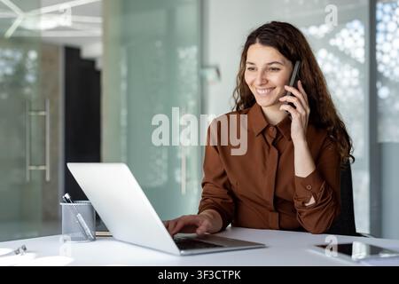 Smiling businesswoman talking on a smartphone and typing on a laptop, managing her work at a modern office desk, embodying efficiency and communication Stock Photo