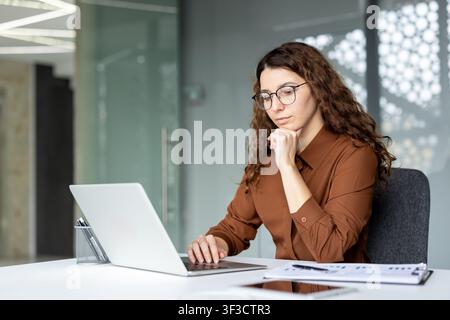 Professional woman in glasses deeply reflecting while using her laptop, analyzing important business data and documents on her desk in a modern corporate office setting Stock Photo