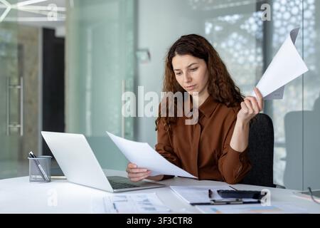Woman concentrating on business documents and financial reports while working with a laptop at a modern corporate office, showing dedication to her accountancy tasks Stock Photo