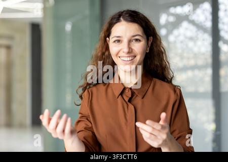 Young professional woman with curly hair smiling and gesturing with her hands while actively communicating and sharing ideas during a business discussion in a contemporary office setting Stock Photo