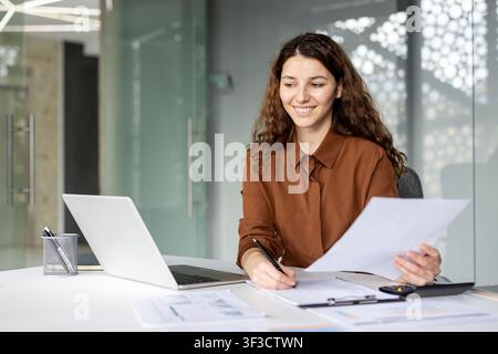 Young businesswoman managing business finances, reviewing financial reports and paperwork, and using a laptop at her desk, representing productivity and corporate success Stock Photo