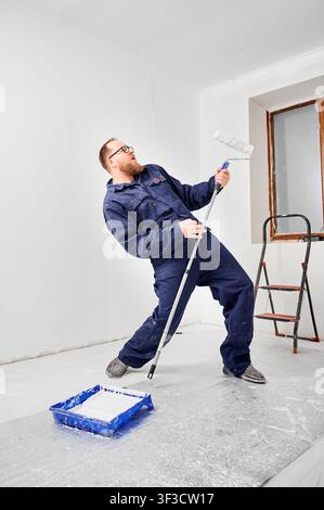 Worker singing and dancing while holding paint roller like guitar in bright room. Fun moment during renovation, positive mood in interior finishing and home improvement concept Stock Photo