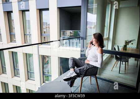 Smiling woman uses smartphone on balcony chair in stylish residential building with large windows. Great for technology promotion, apartment lifestyle, remote work, and city living use Stock Photo