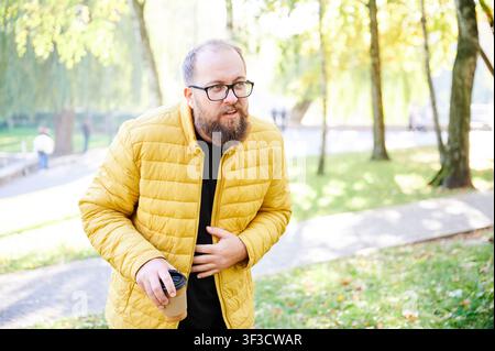 Man experiencing stomach discomfort outdoors. Individual shows distress with hand on abdomen. Man clearly suffering from abdominal pain while standing on garden trail with concerned expression Stock Photo