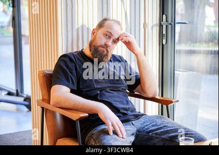 Bearded white man seated in chair, freelance designer taking contemplative break beside window, warm natural light, casual denim, glass of water on table, relaxed posture, reflective atmosphere Stock Photo