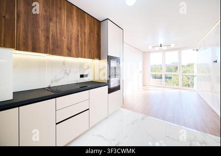 Bright open plan living area with panoramic windows and built in kitchen appliances. Modern apartment interior with wooden floor, marble wall and minimalist luxury design concept Stock Photo