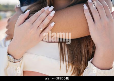 Woman's hands with a diamond engagement ring resting on her fiance's shoulders as they embrace outdoors, a tender close up of love and marriage Stock Photo