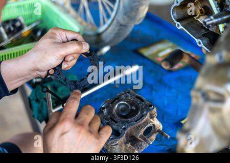 A man holding worn engine gasket over disassembled motorcycle cylinder head on workbench, focused repair scene with tools and greasy parts Stock Photo
