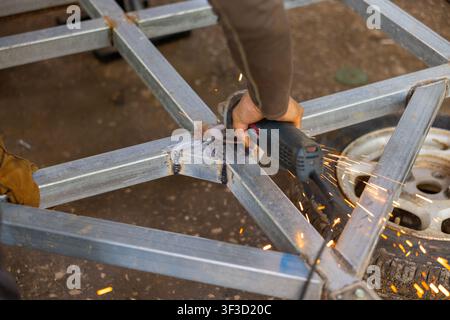 A metalworker using angle grinder to smooth welded steel frame with sparks flying, hands steady and focused in workshop setting Stock Photo